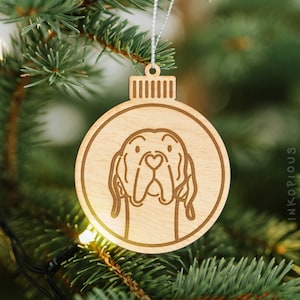 May include: A wooden Christmas ornament shaped like a bauble with a detailed illustration of a Bloodhound dog's head. The ornament is hanging from a white string on a green Christmas tree.