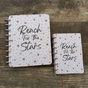 May include: Two white spiral bound notebooks with a gold star and dot pattern. The larger notebook has the text "Reach for the Stars" in black. The smaller notebook has the same text in black.