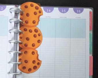 Può includere: Un'agenda con rilegatura argentata e tre fermagli a forma di biscotto. Le graffette sono arancioni con gocce di cioccolato marroni e un morso è stato preso da ognuna. L'agenda è aperta su un calendario di settembre.