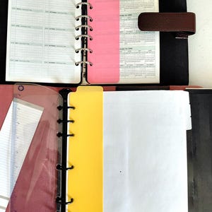 Pu&ograve; includere: Agenda aperto con copertina nera, con un calendario di pianificazione e calendari di riferimento. Divisori rosa e gialli separano le sezioni. L'agenda &egrave; aperta per mostrare le pagine e il raccoglitore ad anelli in metallo.