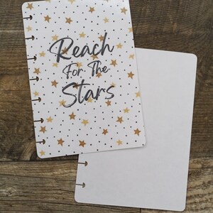 May include: A white and gold planner cover with a gold star pattern and the text "Reach for the Stars". The cover is paired with a blank white planner insert.