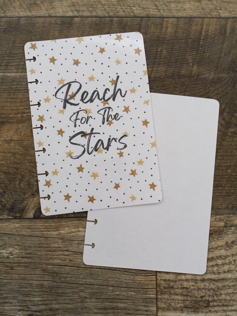 May include: A white and gold planner cover with a gold star pattern and the text "Reach for the Stars". The cover is paired with a blank white planner insert.