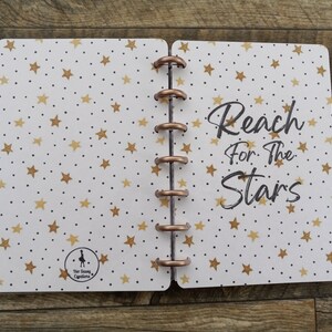 May include: A white planner with a gold star pattern and the text "Reach for the Stars". The planner has a copper ring binding.
