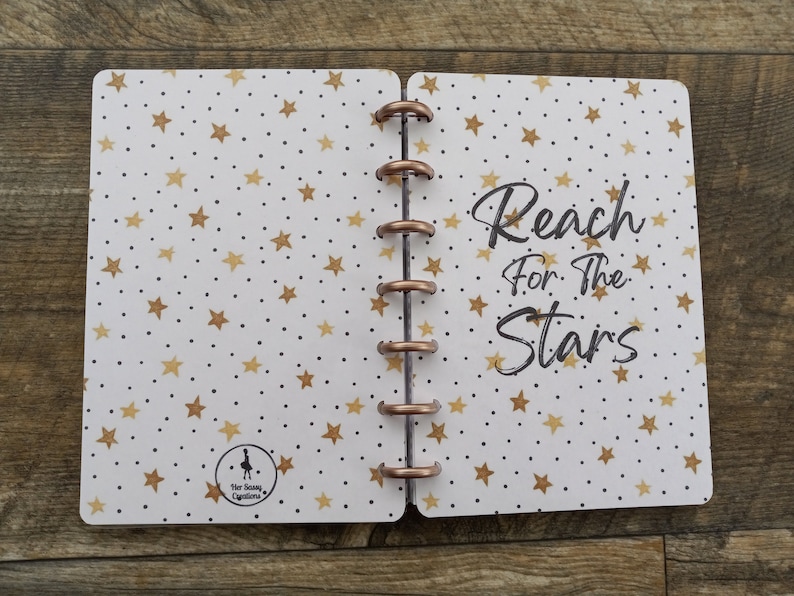 May include: A white planner with a gold star pattern and the text "Reach for the Stars". The planner has a copper ring binding.