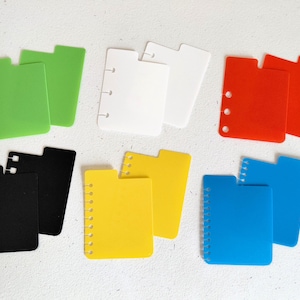 May include: A selection of colourful planner dividers in green, white, red, black, yellow, and blue. Each divider is rectangular with a tab and holes or a spiral binding along the side.