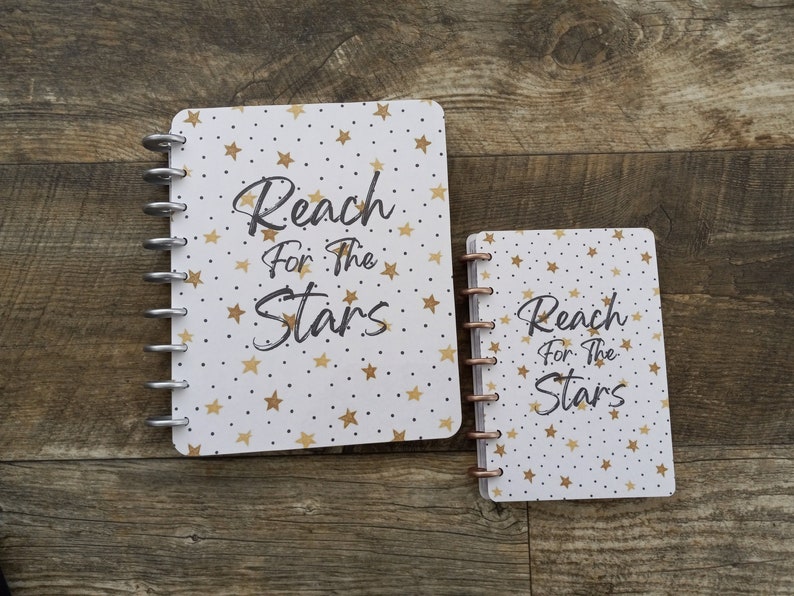 May include: Two white spiral bound notebooks with a gold star and dot pattern. The larger notebook has the text "Reach for the Stars" in black. The smaller notebook has the same text in black.