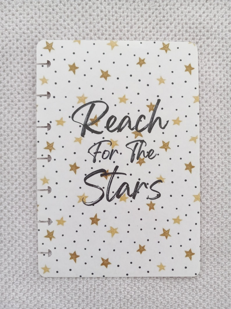 May include: White planner cover with a gold star pattern and the text "Reach for the Stars" in black.