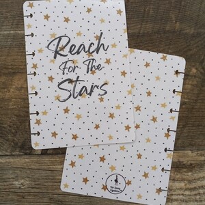 May include: White planner cover with gold stars and black polka dots. The text "Reach for the Stars" is written in black cursive. The bottom right corner has a small black silhouette of a woman with the text "Her Sassy Creations".