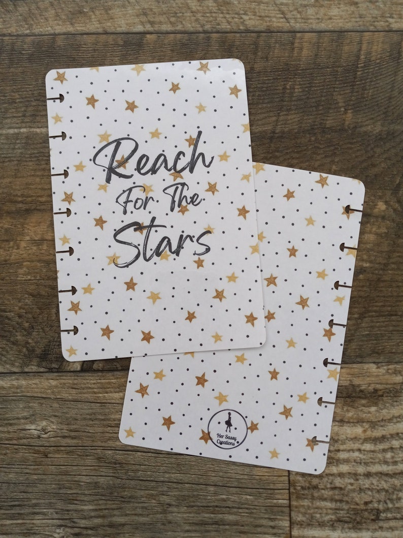 May include: White planner cover with gold stars and black polka dots. The text "Reach for the Stars" is written in black cursive. The bottom right corner has a small black silhouette of a woman with the text "Her Sassy Creations".