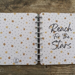 May include: A white notebook with a gold star pattern and black dots. The notebook has a silver coil binding and the text "Reach for the Stars" is written in black on the right side.