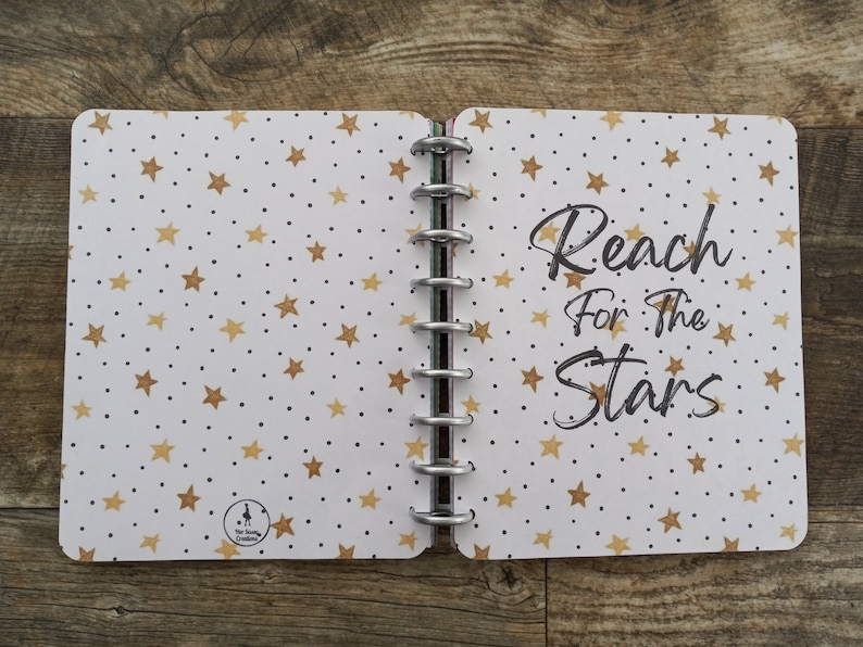 May include: A white notebook with a gold star pattern and black dots. The notebook has a silver coil binding and the text "Reach for the Stars" is written in black on the right side.