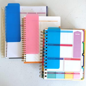 May include: Three spiral-bound planners with colorful covers and dividers. The planners feature blue, pink, and white covers, with gold spiral binding. The pages are organized with daily and weekly layouts, and include sections for to-do lists.