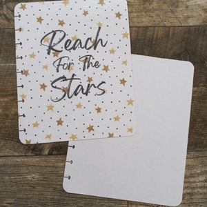 May include: A white planner with a gold star pattern and the text "Reach for the Stars". The planner is open to a blank page.