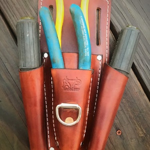 May include: A brown leather tool belt with three pockets. The belt has a metal D-ring and a stitched design. The pockets contain two pairs of pliers and a screwdriver. The pliers have a warning label that reads "WARNING: NOT INSULATED.  WILL NOT PROTECT AGAINST ELECTRICAL SHOCK."