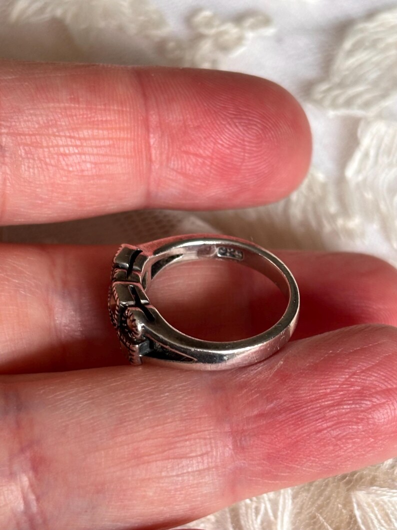 Pu&ograve; includere: Un anello d'argento con un design decorativo. L'anello ha una fascia liscia e lucida e una parte superiore dettagliata con un motivo strutturato. L'anello &egrave; tenuto tra le dita.