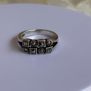 Pu&ograve; includere: Un anello d'argento con una serie di elementi decorativi quadrati. Ogni quadrato &egrave; adornato con pietre pi&ugrave; piccole e scure, creando un motivo strutturato. L'anello &egrave; posto su una superficie bianca, mettendo in risalto il suo design.