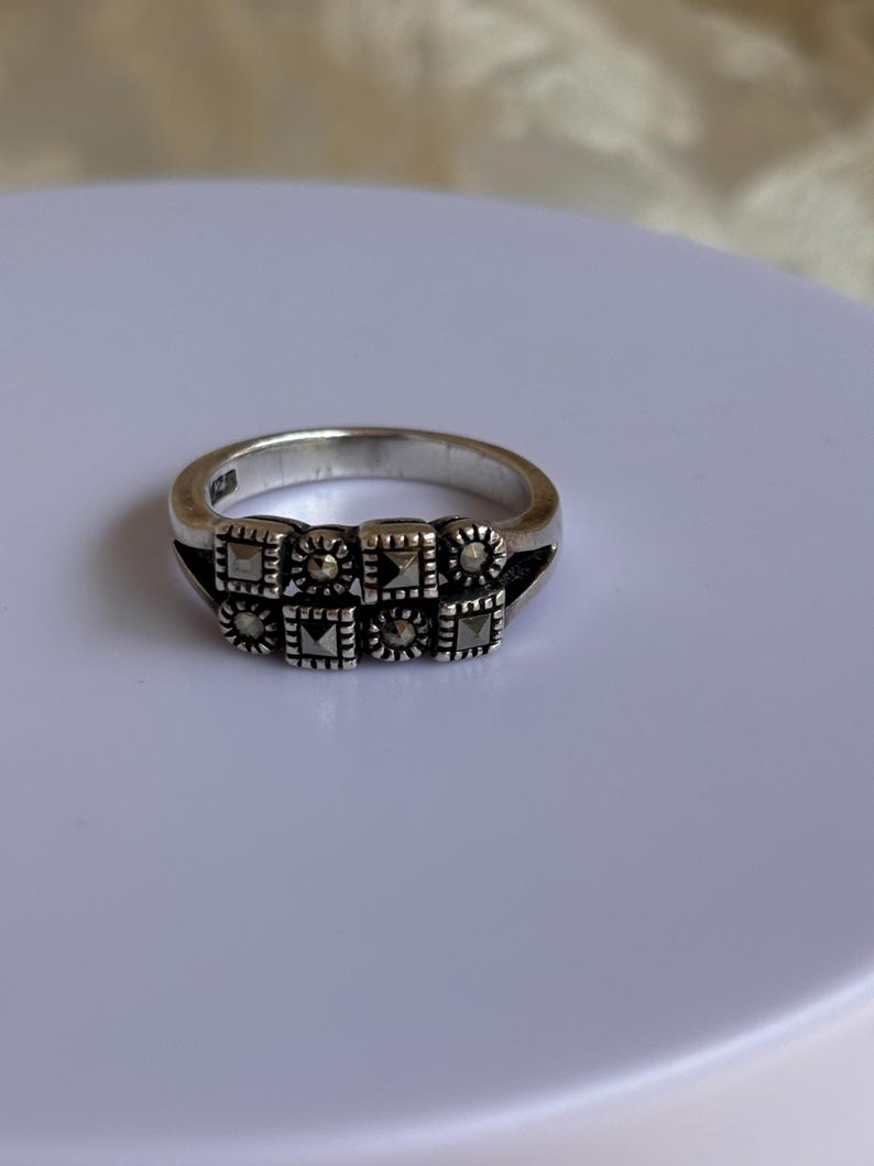 Pu&ograve; includere: Un anello d'argento con una serie di elementi decorativi quadrati. Ogni quadrato &egrave; adornato con pietre pi&ugrave; piccole e scure, creando un motivo strutturato. L'anello &egrave; posto su una superficie bianca, mettendo in risalto il suo design.