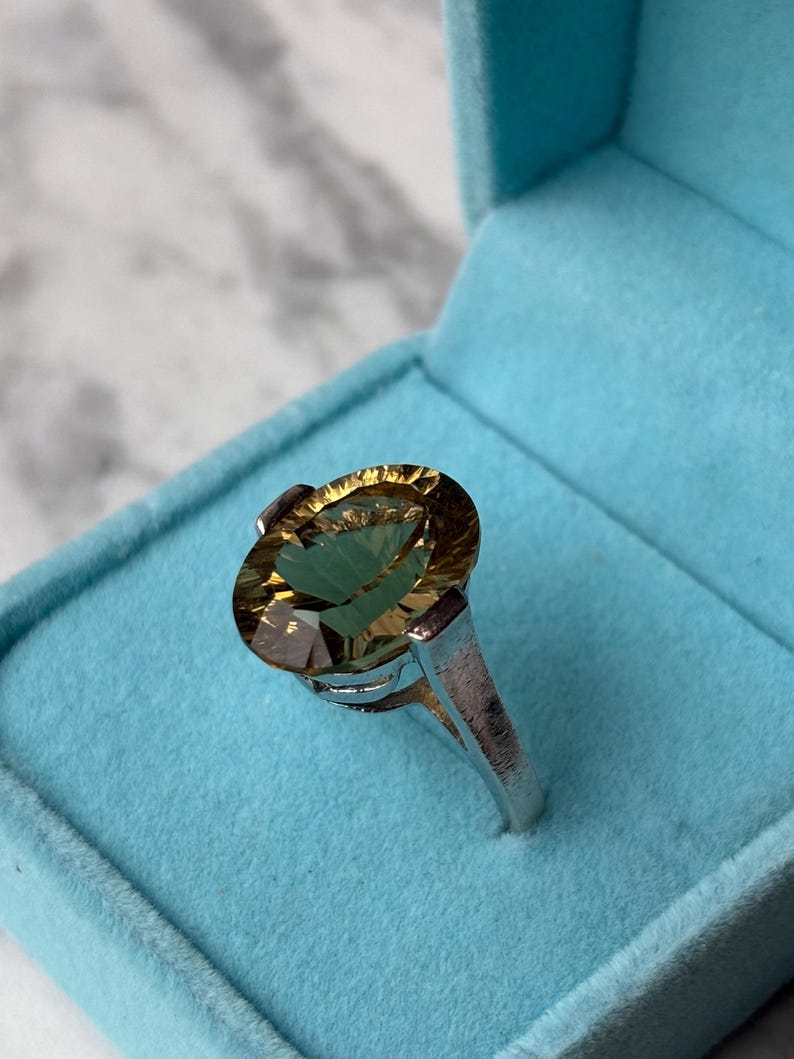 Pu&ograve; includere: Un anello con una grande pietra preziosa dal taglio ovale color marrone dorato, incastonata in una fascia di metallo color argento lucido. La pietra sfaccettata mostra tonalit&agrave; ambra e miele ed &egrave; presentata in una scatola blu brillante.