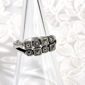 Op de afbeelding: Een zilveren ring met een dubbel bandontwerp. De ring heeft vierkante en ronde markasietstenen in een geometrisch patroon. De ring wordt gepresenteerd op een heldere, glazen kegelvormige standaard tegen een witte achtergrond.
