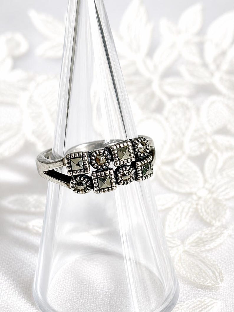 Pu&ograve; includere: Un anello d'argento con un design a doppia fascia. L'anello presenta pietre di marcasite quadrate e rotonde disposte in un motivo geometrico. L'anello &egrave; esposto su un supporto conico di vetro trasparente su sfondo bianco.