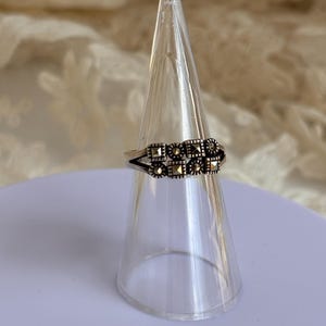 Pu&ograve; includere: Un anello d'argento con un design rettangolare con piccole pietre scure. L'anello &egrave; esposto su un supporto trasparente a forma di cono, su uno sfondo morbido e neutro. Il design dell'anello &egrave; simmetrico e dettagliato.
