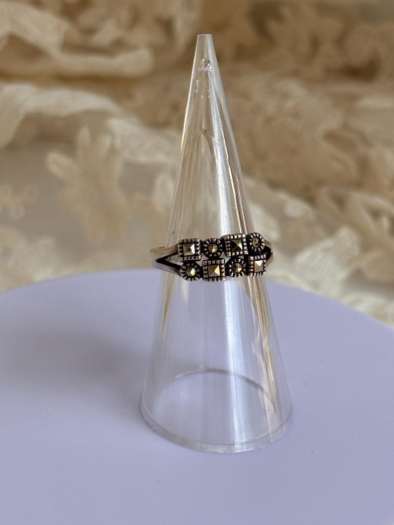 Pu&ograve; includere: Un anello d'argento con un design rettangolare con piccole pietre scure. L'anello &egrave; esposto su un supporto trasparente a forma di cono, su uno sfondo morbido e neutro. Il design dell'anello &egrave; simmetrico e dettagliato.