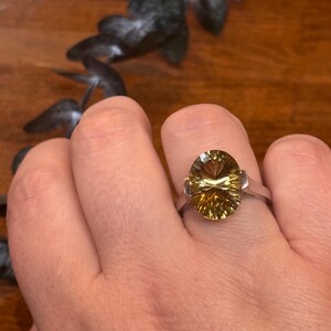 Pu&ograve; includere: Un anello color argento con una grande pietra preziosa giallo oro, taglio ovale e superficie sfaccettata. La pietra &egrave; incastonata in una semplice fascia metallica che la sostiene sui lati, conferendo un aspetto luminoso e trasparente.