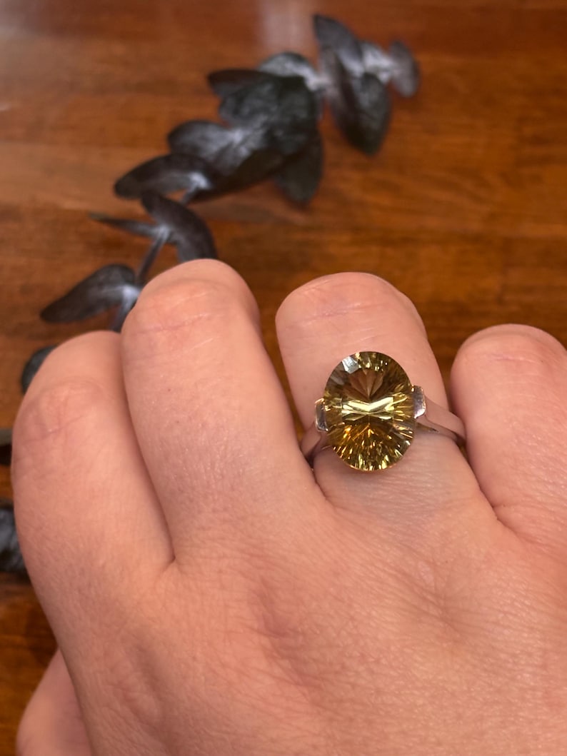 Pu&ograve; includere: Un anello color argento con una grande pietra preziosa giallo oro, taglio ovale e superficie sfaccettata. La pietra &egrave; incastonata in una semplice fascia metallica che la sostiene sui lati, conferendo un aspetto luminoso e trasparente.