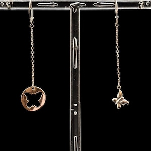 May include: A pair of mismatched drop earrings featuring a silver-toned metal chain. One earring displays a circular charm with a butterfly cutout, while the other earring features a small, three-dimensional butterfly charm dangling from the chain.