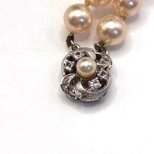 May include: A strand of round, light peach-colored pearls connected to a square, silver-toned metal clasp. The clasp features a central pearl surrounded by four small, clear, faceted stones set within a textured, swirling metal frame design.