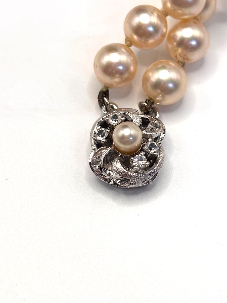 May include: A strand of round, light peach-colored pearls connected to a square, silver-toned metal clasp. The clasp features a central pearl surrounded by four small, clear, faceted stones set within a textured, swirling metal frame design.
