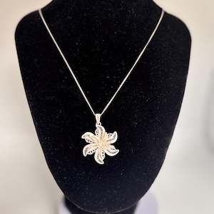 Puede incluir: Un collar con colgante de flor de filigrana plateada expuesto sobre un busto de terciopelo negro. El colgante es un diseño calado detallado en forma de flor de seis pétalos. La cadena del collar es delicada y plateada.