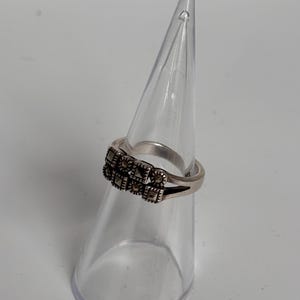 Pu&ograve; includere: Un anello d'argento con un design rettangolare di piccole pietre scure. L'anello &egrave; esposto su un supporto trasparente a forma di cono, su sfondo bianco. La fascia dell'anello &egrave; sottile e semplice, in contrasto con l'incastonatura dettagliata delle pietre.