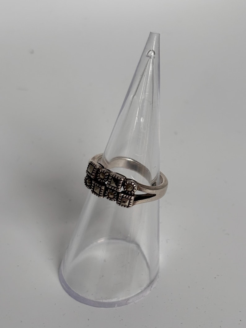 Pu&ograve; includere: Un anello d'argento con un design rettangolare di piccole pietre scure. L'anello &egrave; esposto su un supporto trasparente a forma di cono, su sfondo bianco. La fascia dell'anello &egrave; sottile e semplice, in contrasto con l'incastonatura dettagliata delle pietre.
