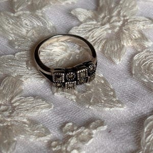 Pu&ograve; includere: Un anello d'argento con un design rettangolare. L'anello presenta una serie di piccole pietre quadrate disposte in fila sulla parte superiore. L'anello &egrave; posto su uno sfondo bianco strutturato.