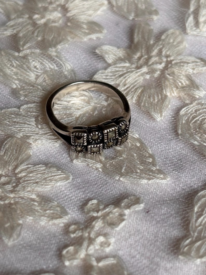 Pu&ograve; includere: Un anello d'argento con un design rettangolare. L'anello presenta una serie di piccole pietre quadrate disposte in fila sulla parte superiore. L'anello &egrave; posto su uno sfondo bianco strutturato.