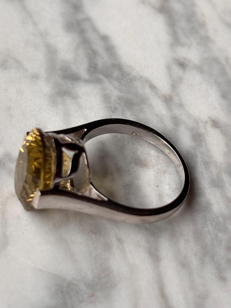 Pu&ograve; includere: Un anello color argento con una grande pietra preziosa gialla a taglio ovale montata su una griffa. L'interno della fascia dell'anello &egrave; chiaramente impresso con il marchio 925, che indica una composizione in argento sterling. Il design presenta una finitura metallica lucida.