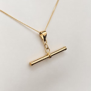 May include: A gold necklace featuring a T-bar pendant suspended from a delicate chain. The T-bar and the pendant bail are both clearly stamped with the number 375, indicating the gold purity. The metal has a polished, reflective finish.