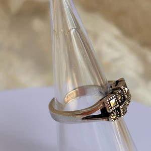 Pu&ograve; includere: Un anello d'argento con un design quadrato e ornato con piccole pietre scure. L'anello &egrave; esposto su un supporto conico trasparente, mettendo in risalto i suoi dettagli intricati e lo stile vintage.