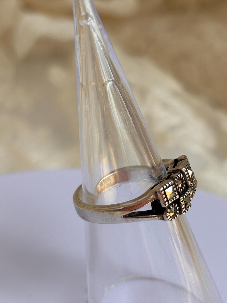 Pu&ograve; includere: Un anello d'argento con un design quadrato e ornato con piccole pietre scure. L'anello &egrave; esposto su un supporto conico trasparente, mettendo in risalto i suoi dettagli intricati e lo stile vintage.