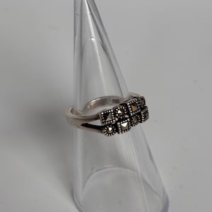 Pu&ograve; includere: Un anello d'argento con un gruppo di piccole pietre quadrate. L'anello &egrave; esposto su un supporto trasparente a forma di cono su sfondo bianco. Le pietre sono scure, forse marcasite, disposte in un motivo geometrico.
