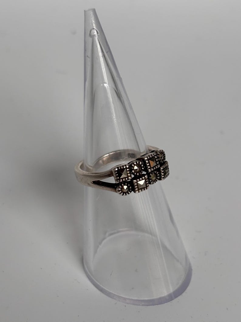 Pu&ograve; includere: Un anello d'argento con un gruppo di piccole pietre quadrate. L'anello &egrave; esposto su un supporto trasparente a forma di cono su sfondo bianco. Le pietre sono scure, forse marcasite, disposte in un motivo geometrico.