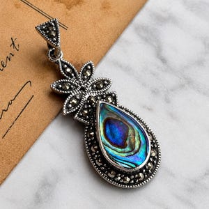 Puede incluir: Colgante plateado con incrustación de concha de abulón en forma de lágrima. El colgante presenta un diseño floral con detalles de marcasita y una parte superior triangular. La concha iridiscente muestra tonos de azul, verde y morado.