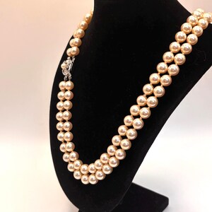 May include: A double-strand necklace featuring round, cream-colored pearls with a soft luster. The necklace is secured with a silver-toned metal clasp that is embellished with a small, central pearl and clear, sparkling accent stones.