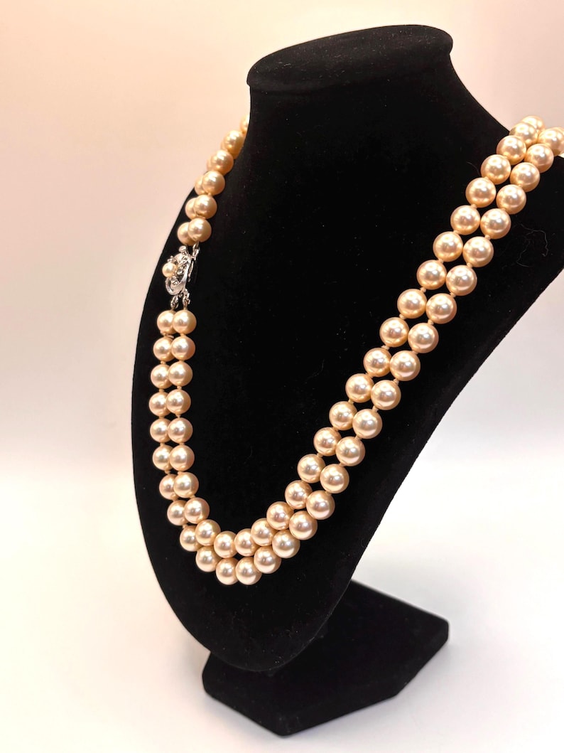 May include: A double-strand necklace featuring round, cream-colored pearls with a soft luster. The necklace is secured with a silver-toned metal clasp that is embellished with a small, central pearl and clear, sparkling accent stones.