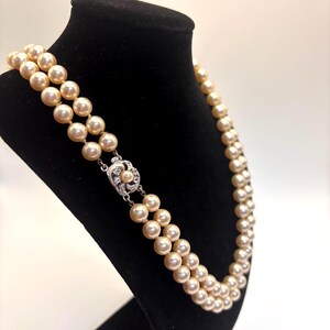 May include: A double-strand necklace featuring cream-colored pearls of uniform size. The necklace is secured with a silver-toned metal clasp that is circular in shape and decorated with small, clear, sparkling stones and a single central pearl.