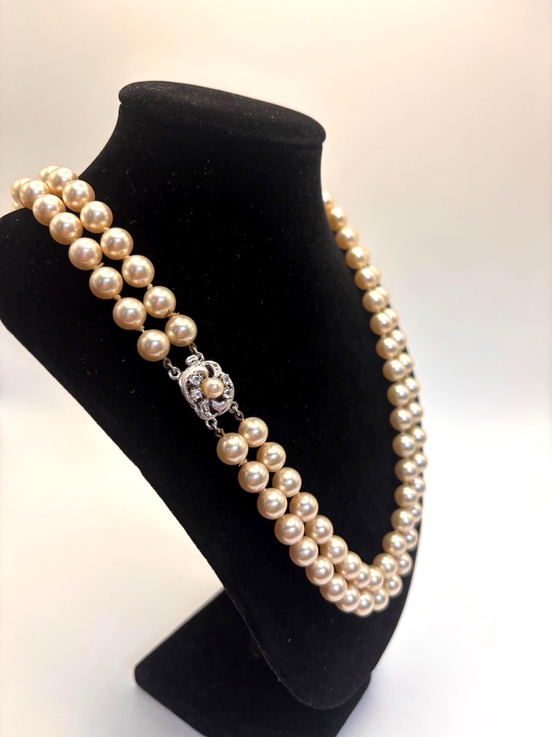 May include: A double-strand necklace featuring cream-colored pearls of uniform size. The necklace is secured with a silver-toned metal clasp that is circular in shape and decorated with small, clear, sparkling stones and a single central pearl.