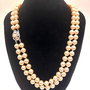 May include: A double-strand necklace featuring round, cream-colored pearls of uniform size. The necklace is secured with a silver-toned metal clasp that is embellished with small, clear, sparkling stones and a single central pearl accent.
