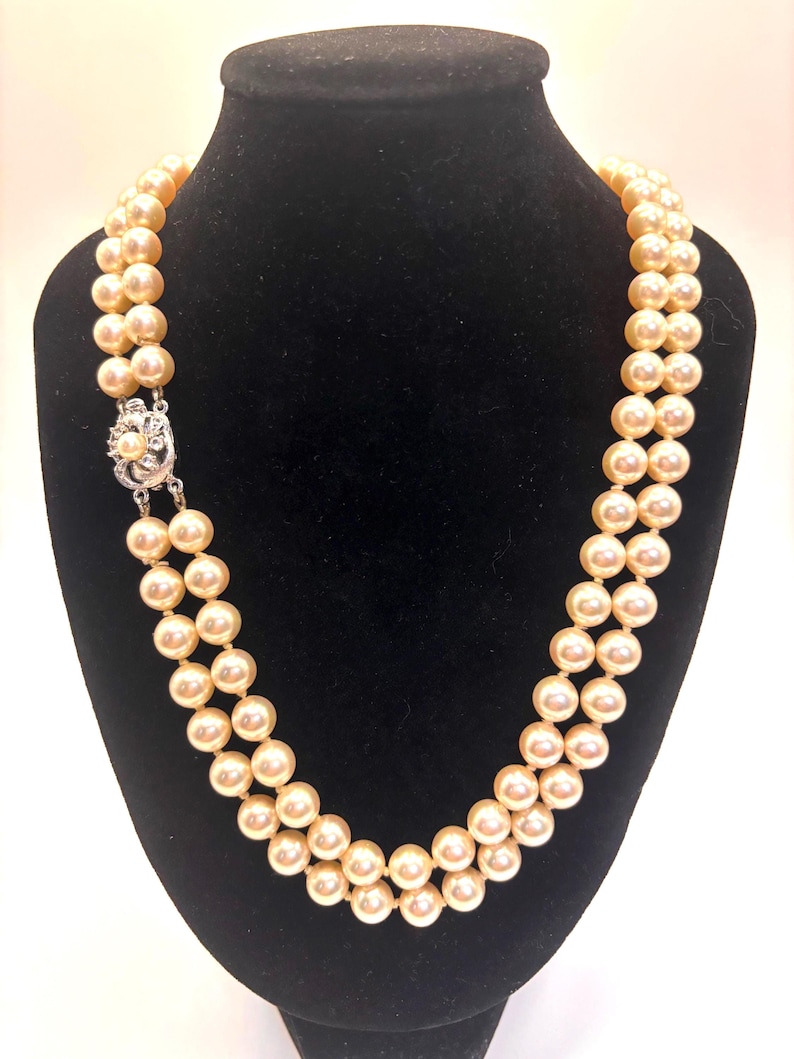 May include: A double-strand necklace featuring round, cream-colored pearls of uniform size. The necklace is secured with a silver-toned metal clasp that is embellished with small, clear, sparkling stones and a single central pearl accent.