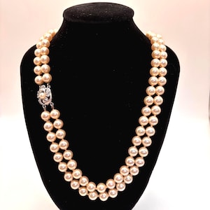 May include: A double-strand necklace featuring round, cream-colored pearls. The two strands are connected by a silver-toned metal clasp shaped like a flower, which is accented with small, clear, sparkling stones and a single central pearl.